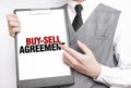 BUY-SELL AGREEMENT inscription on a notebook in the hands of a businessman on a gray background, a man points with a finger to the