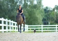 Smiling woman galloping on horse while her puppy chases after them around arena.