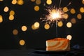 Cake with burning sparkler on black table against blurred festive lights. Space for text Royalty Free Stock Photo