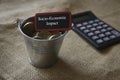 A calculator, small silver steel bucket with gold coins and red signboard written with Socio-Economic Impact. Business and finance