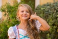 Call me sign. Cute little blonde girl child shows by gesture that she speaks on the phone smiling toothless isolated green shrubs