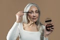 Calm aged european lady with gray hair applies shadows with brush on her eyes, shows cosmetics