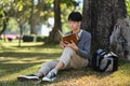 Calm asian man reading book while sitting under tree on green grass at sunny beautiful day with sunlight Royalty Free Stock Photo