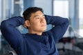 A calm Asian man sits in the office on a chair, leans back, hands behind his head. Resting Royalty Free Stock Photo