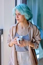 Calm caucasian female with unusual coloured blue hair on near window with mug