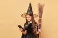 Calm cute little girl in black cloak clothing with broom isolated over beige background using mobile phone browsing internet