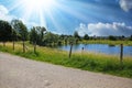 Calm cycling track in beautiful rural dutch landscape, small lake on farm land, green meadows, blue summer sky with sun rays - Royalty Free Stock Photo