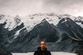 calm disheveled young caucasian man bundled up in black coat in front of huge snowy big rocky mountains over calm lake