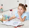 Calm dreaming kid girl is lying on soft carpet drawing coloring pictures of dolls with colorful pencils