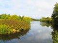 The calm, flat waters of a lake twists between banks covered with bushes and trees.