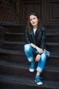A calm girl in sneakers, jeans and a leather jacket sits on wood