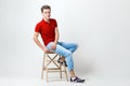 Calm handsome dark-haired guy wearing red shirt and blue jeans sitting on wooden stool over white background Royalty Free Stock Photo