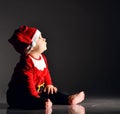 Calm infant boy toddler in santa claus costume is sitting barefooted on ice looking up carefully in corner at copy space