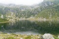 Calm lake in mountains with clear water, with reflection of rocky mountains on surface on foggy day