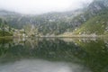 Calm lake in mountains with clear water, with reflection of rocky mountains on surface on foggy day Royalty Free Stock Photo