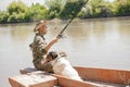 Calm male fisher throwing hook into water, while sitting in rowing boat with dog in hot day. Royalty Free Stock Photo