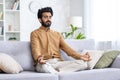 Calm middle eastern man sitting on pillowed sofa in siddhasana position and using hands mudra with closed eyes. Calm guy