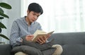 Calm millennial asian man smelling on couch and reading book, spending quality time at home