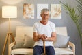 Calm relaxed man pensioner wearing white T-shirt and jeans sitting on sofa in living room holding walking cane looking at camera Royalty Free Stock Photo