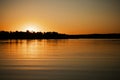 Calm rippled lake with sun almost set below horizon covering surface of water with its rays with trees on shore in