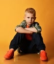 Blond schoolboy teenager, regular guy in checkered shirt, orange t-shirt and jeans sits on floor looking at camera Royalty Free Stock Photo