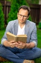 Calm unshaven man in glasses sits on grass reading book outdoors on summer day Royalty Free Stock Photo