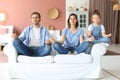 Calm young family with little daughter sit on couch practice yoga together, happy parents with small preschooler girl