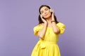 Calmed young woman in yellow dress keeping eyes closed, listen music, putting hands on headphones isolated on pastel Royalty Free Stock Photo