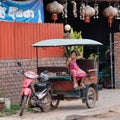 A little Asian girl sits in a moto rickshaw near a house with red lanterns