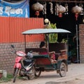A little Asian girl sits in a moto rickshaw near a house with red lanterns