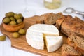 Camembert cheese, sliced fresh baguette, olives on a wooden board on the table. A wineglass and a bottle of white wine blurred in Royalty Free Stock Photo