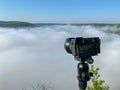 A camera with a tripod at sunrise looks at a foggy canyon.