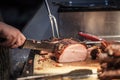 Canadian bacon, also known as peameal, for sale, grilled and cooked, being cut wuith a knife on a wooden plank, surrounded by othe