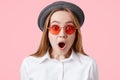 Candid shot of amazed terrified female with wide opened mouth, dressed in white shirt, black stylish hat, poses against pink backg