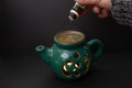 Candle essential oil burner in shape of a green tea pot, with a hand pouring a small bottle on dark background, soft focus close