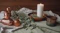a candle and some copper containers on a table with a white cloth and a green leafy branch in the middle of the table, and a few