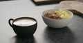 Cappuccino in black cup with granola with mango in white bowl on kitchen