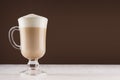 Cappuccino coffee in elegant glass with foam on white table and dark brown wall, copy space.