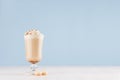 Cappuccino coffee with whipped cream, cocoa powder and biscuits in transparent glass on pastel blue wall and white wood board.