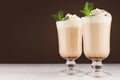 Cappuccino coffee with whipped cream, green mint, cocoa powder in two transparent glass on dark brown wall and white wood board. Royalty Free Stock Photo