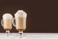 Cappuccino coffee with whipped cream in two transparent glass with handles on dark brown wall and white wood board. Royalty Free Stock Photo