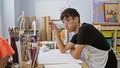 Captivating glimpse into a young hispanic man\'s world, artist, brush in hand, lost in thought while drawing at his studio