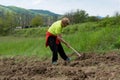 A woman with a hoe is breaking up the soil and creating rows and holes for planting potatoes on a sunny and warm spring day