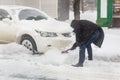 Car blocked with snow drift on city street. Man cleaning vehicle from snow with brush during heavy snowfall, blizzard and storm.