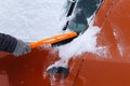 Car covered with snow. Brush in mans hand. Man in gray gloves is brushing orange car from snow