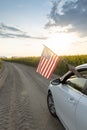 car drives along country road against background of setting sun, blooming rapeseed field and American flag from window Royalty Free Stock Photo