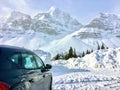 A car parked overlooking incredible view of a dark green forest in the foreground and snow covered mountains peaks