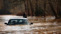 A car submerged up to its hood in floodwater with a person standing on top trying to push it out of harms way