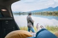 Car trunk view with woman legs in white sneakers on the man dressed warm knitted clothes and jeans enjoying the mountain lake view Royalty Free Stock Photo