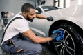 Car washing and detailing photo. African man worker in protective overalls and rubber gloves, washing car alloy wheel on